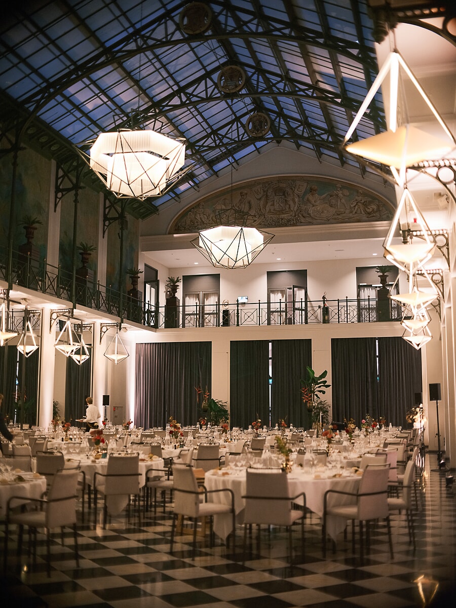 Grote zaal met glazen tongewelf en beschilderde wapenschilden, ronde tafels gedekt onder geometrische hanglampen in Amsterdam