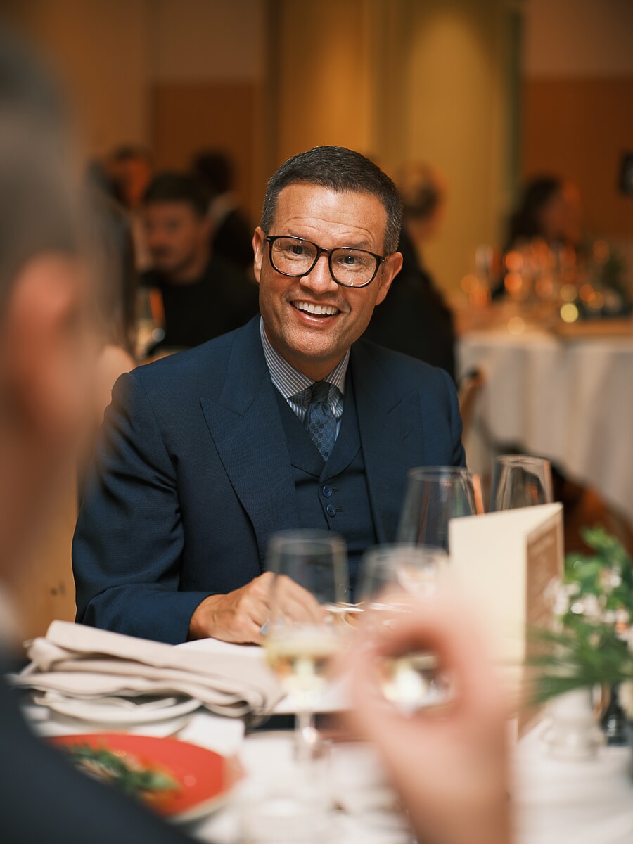 Gast in donkerblauw driedelig pak lacht aan de dinertafel, wijnglazen vangen het licht voor hem, eventfotografie in Amsterdam