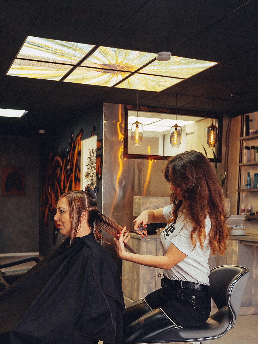 Kapper verdeelt het natte haar van een klant in een salon met een bedrukt lichtplafond en hanglampen, brandingfotografie in Amsterdam