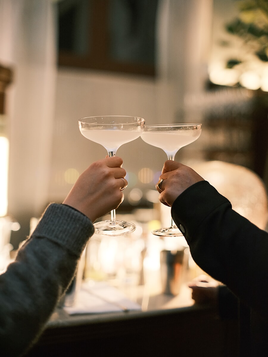 Twee handen klinken met coupeglazen in warm strijklicht, met kaarsen onscherp erachter