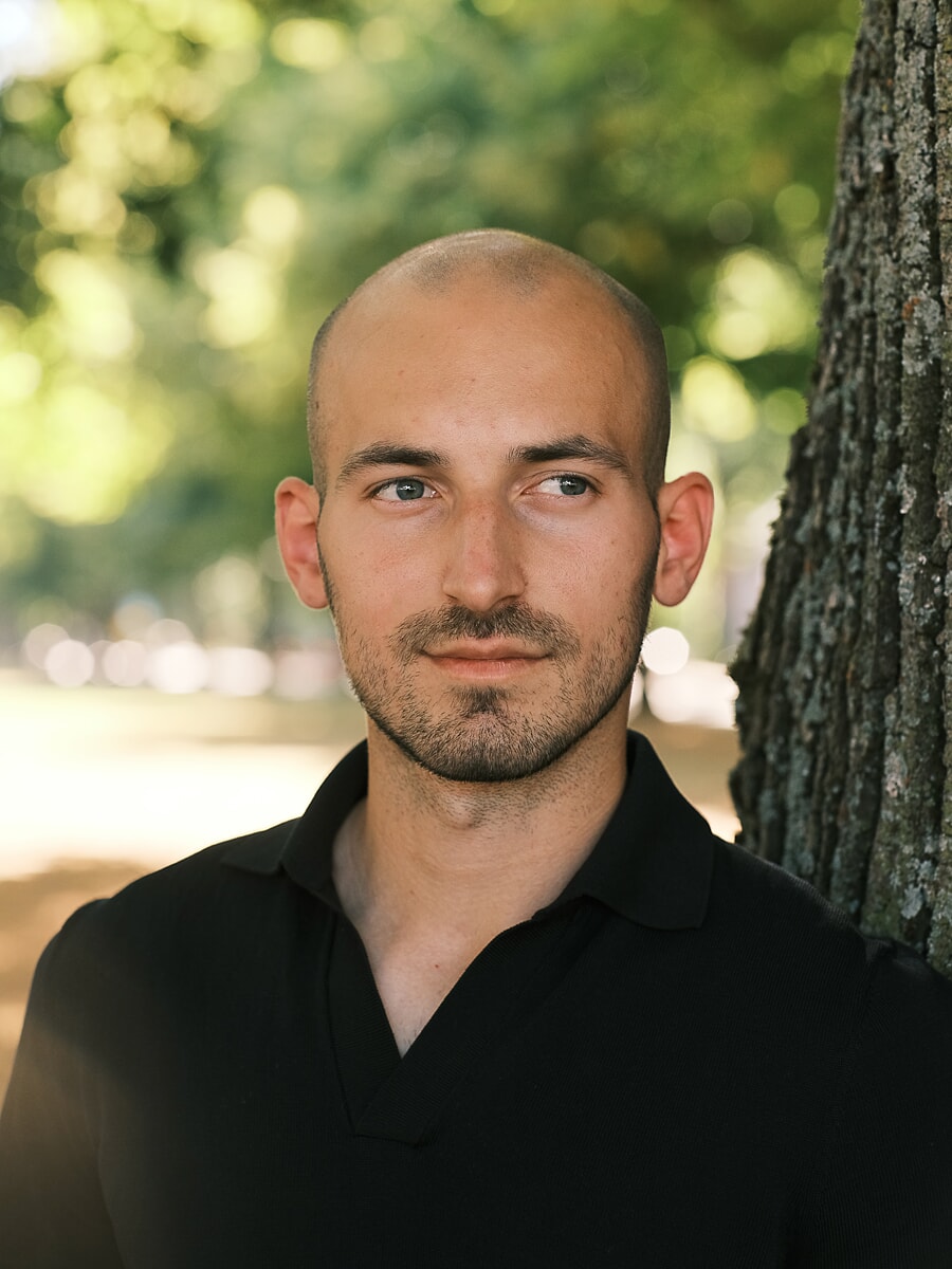 Portret van hoofd en schouders van een man in een zwarte polo naast een boomstam, met groene zomerse bokeh erachter