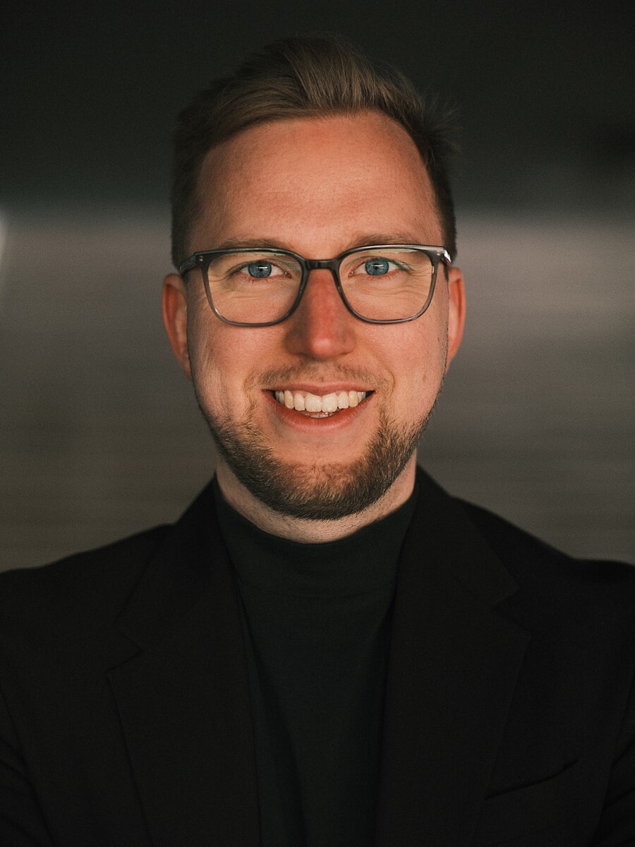 Headshot van dichtbij van een man met bril in een zwarte blazer over een coltrui tegen een donkere achtergrond