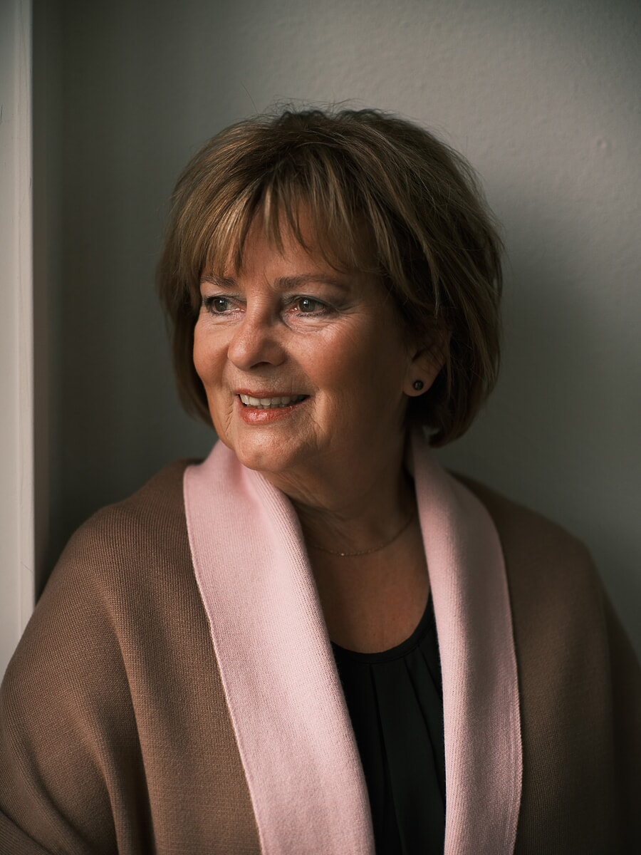 Vrouw in een kameelkleurige omslagdoek met roze bies kijkt in zacht raamlicht, een zakelijk headshotportret in Amsterdam