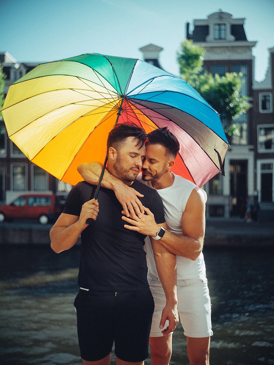 Twee mannen omhelzen elkaar onder een regenboogparaplu langs een Amsterdamse gracht, een levendig koppelportret