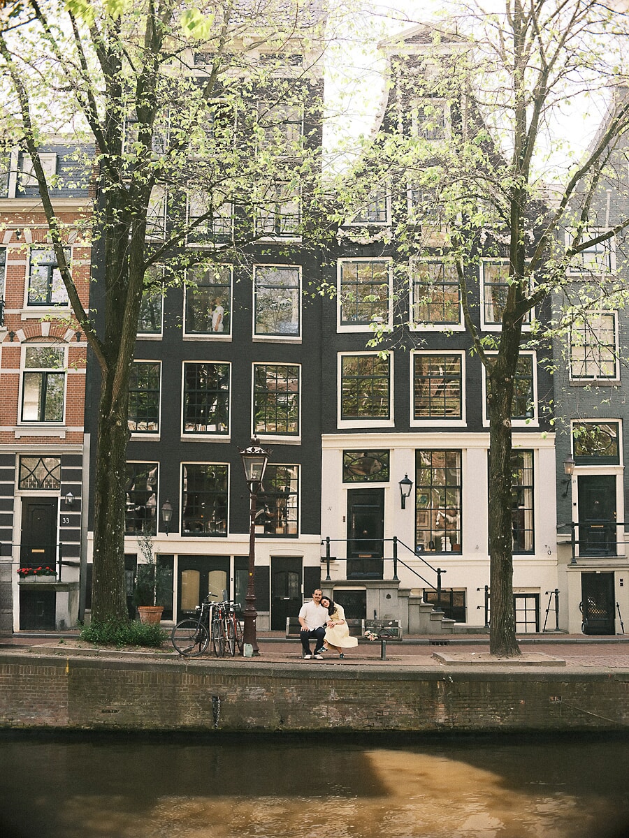 Stel op een bankje onder voorjaarsbomen voor Amsterdamse grachtenpanden, breed in beeld tijdens hun loveshoot