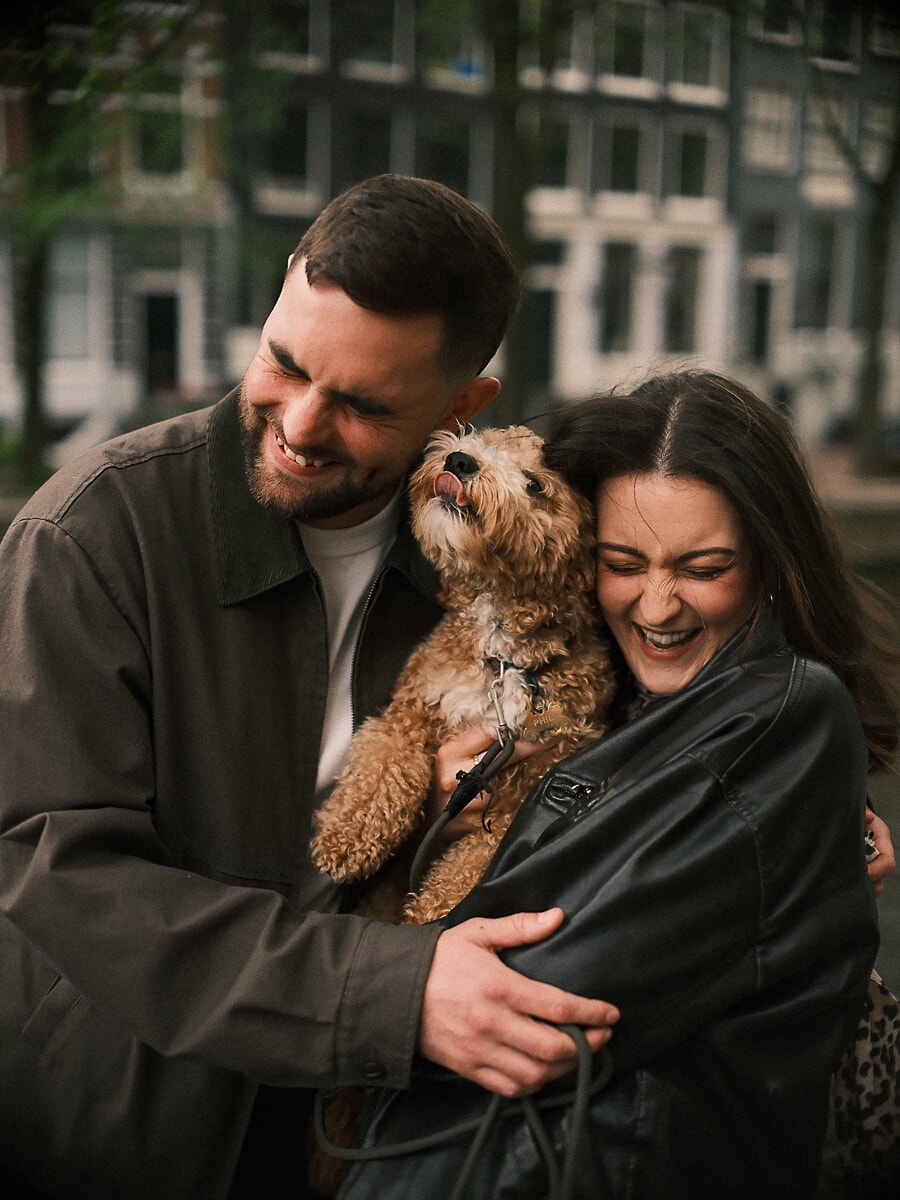 Stel lacht met hun kleine hond in een straat langs een Amsterdamse gracht tijdens hun loveshoot