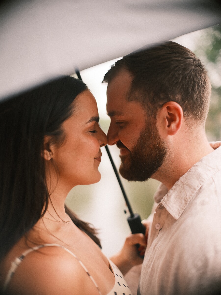 Stel neus tegen neus onder een paraplu in de regen, een intiem koppelportret uit hun loveshoot in Amsterdam