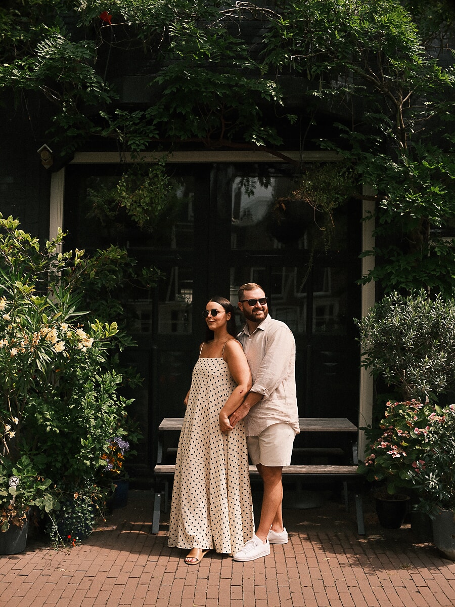 Stel leunt tegen elkaar aan onder klimgroen voor een donkere Amsterdamse gevel, een ontspannen loveshoot-portret