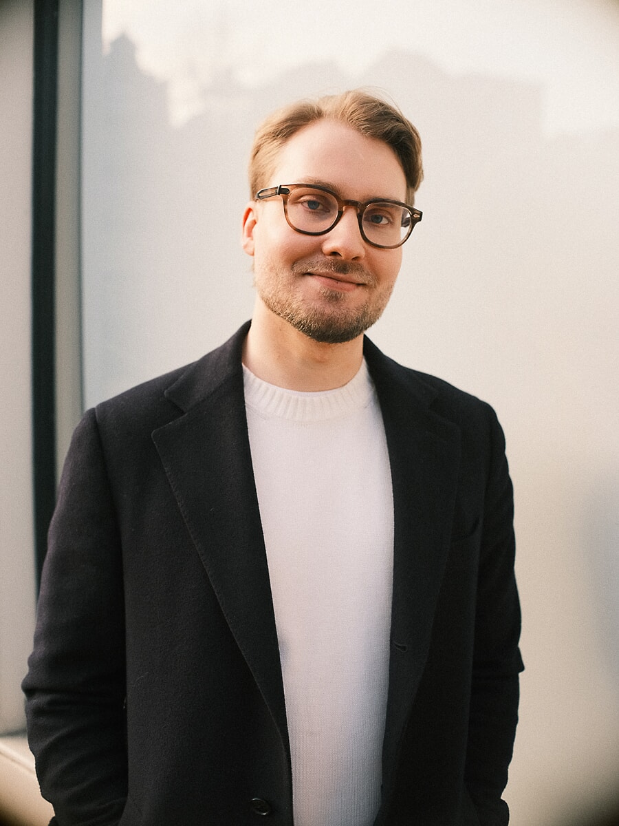 Portret van hoofd en schouders van een man met bril in zwarte jas over een witte trui tegen een lichte muur, voor een online datingprofiel