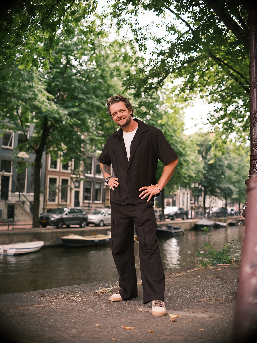 Man staat ten voeten uit met een hand in zijn zij, in zwart overhemd en wijde broek langs een gracht, een ontspannen confidenceportret