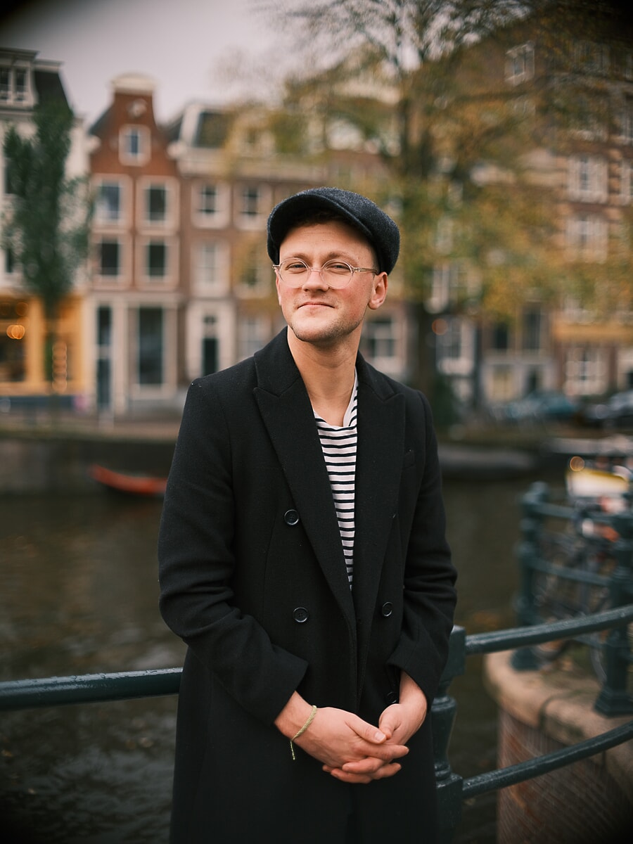 Man met flatcap en ronde bril, handen gevouwen, zwarte jas over een gestreept shirt, een portret langs de gracht