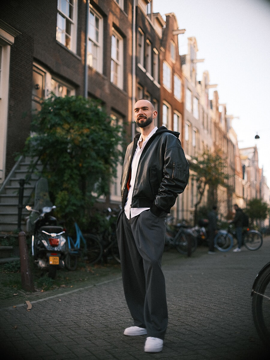 Man in zwart leren bomberjack en wijde grijze broek in een straat langs een gracht in laag herfstlicht, een portret ten voeten uit