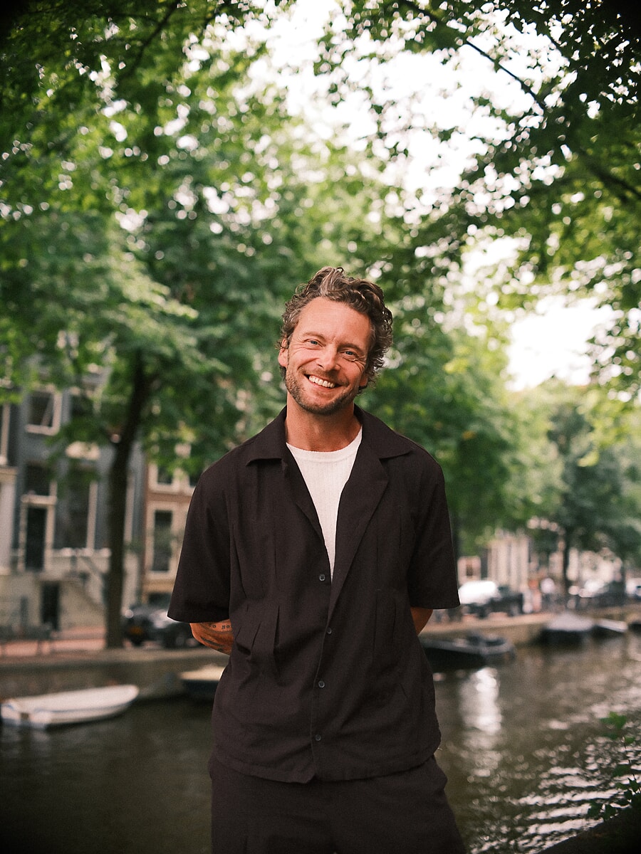 Man met krullend haar lacht in een zwart overhemd langs een gracht vol afgemeerde boten, een zomers portret in Amsterdam