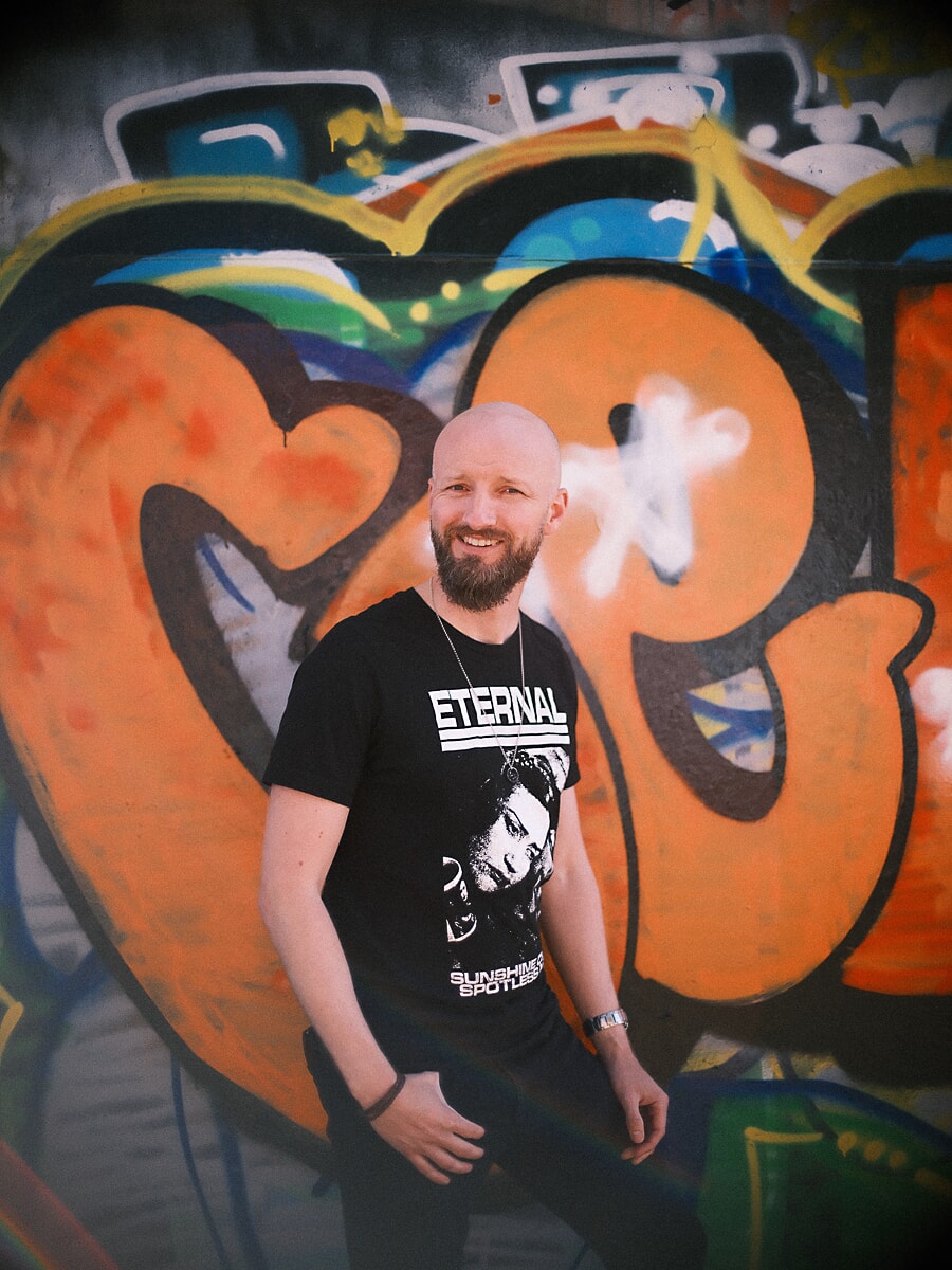 Man met baard lacht voor een groot oranje graffitikunstwerk, een kleurrijk beeld uit zijn datingshoot