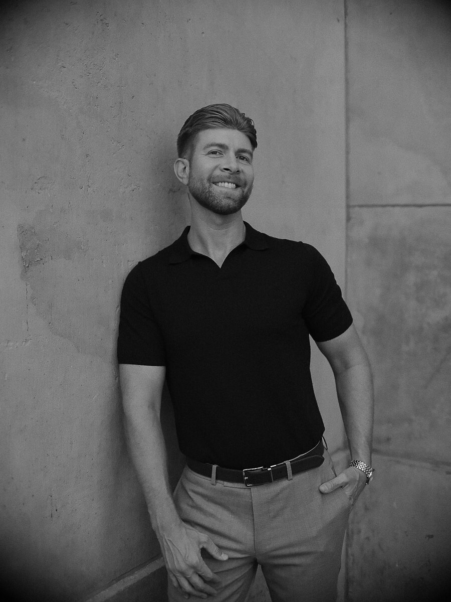 Man leunt met één hand in zijn zak tegen een betonnen muur, een ontspannen zwart-wit portret