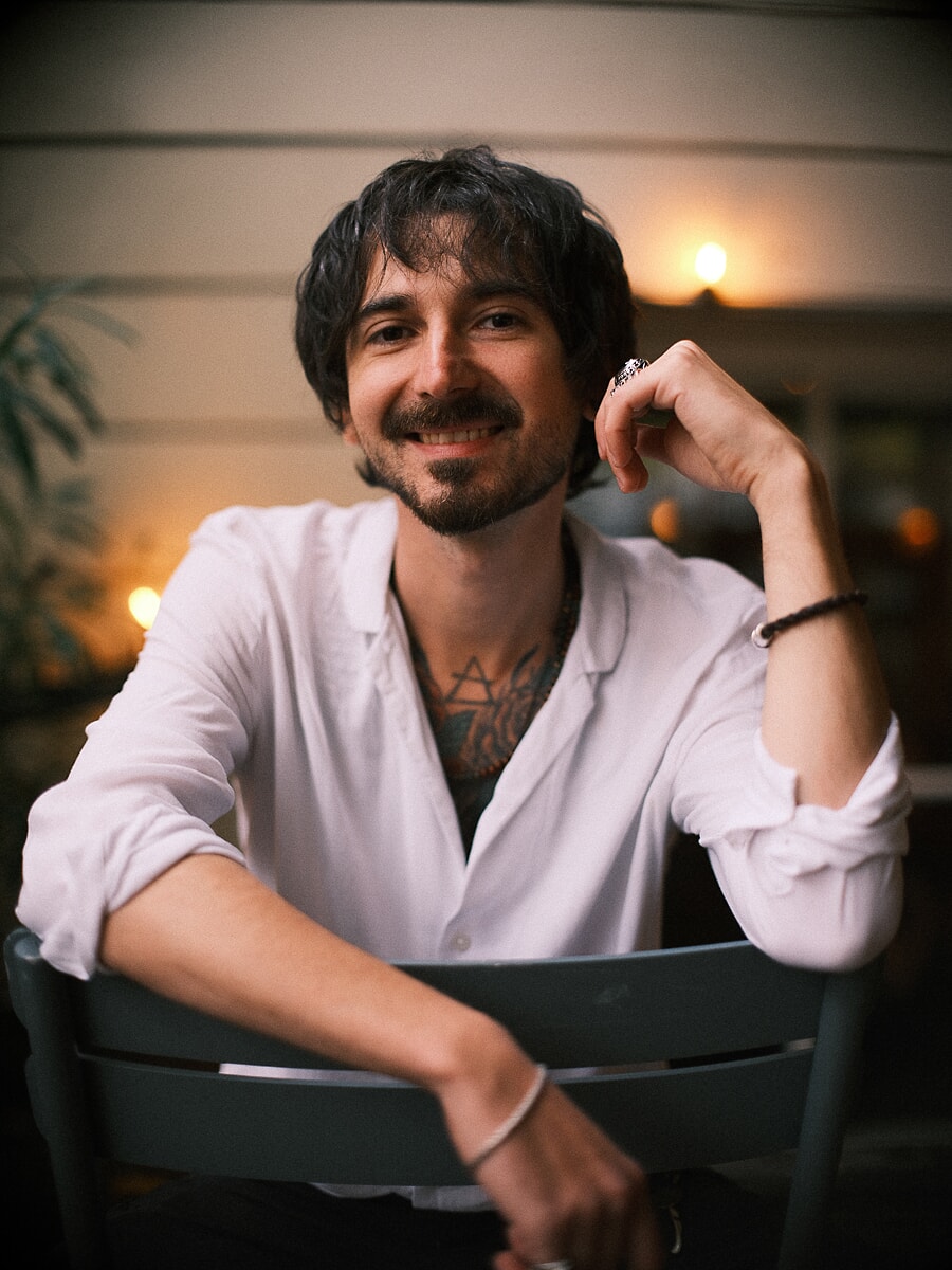 Man in open wit overhemd op een caféterras in de avond, met warme lichtjes onscherp erachter, een avondlijk datingportret