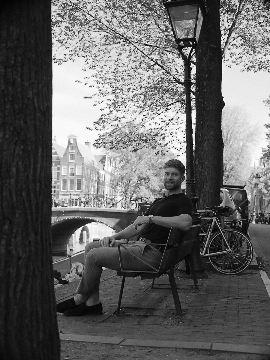 Man in donkere polo op een bankje onder de bomen langs een Amsterdamse gracht, een zwart-wit portret