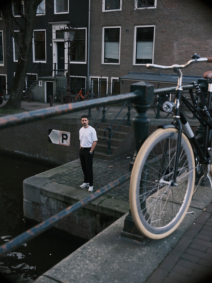 Man in wit T-shirt op een kade langs de gracht, gezien langs een fietswiel en reling, een documentair portret in Amsterdam