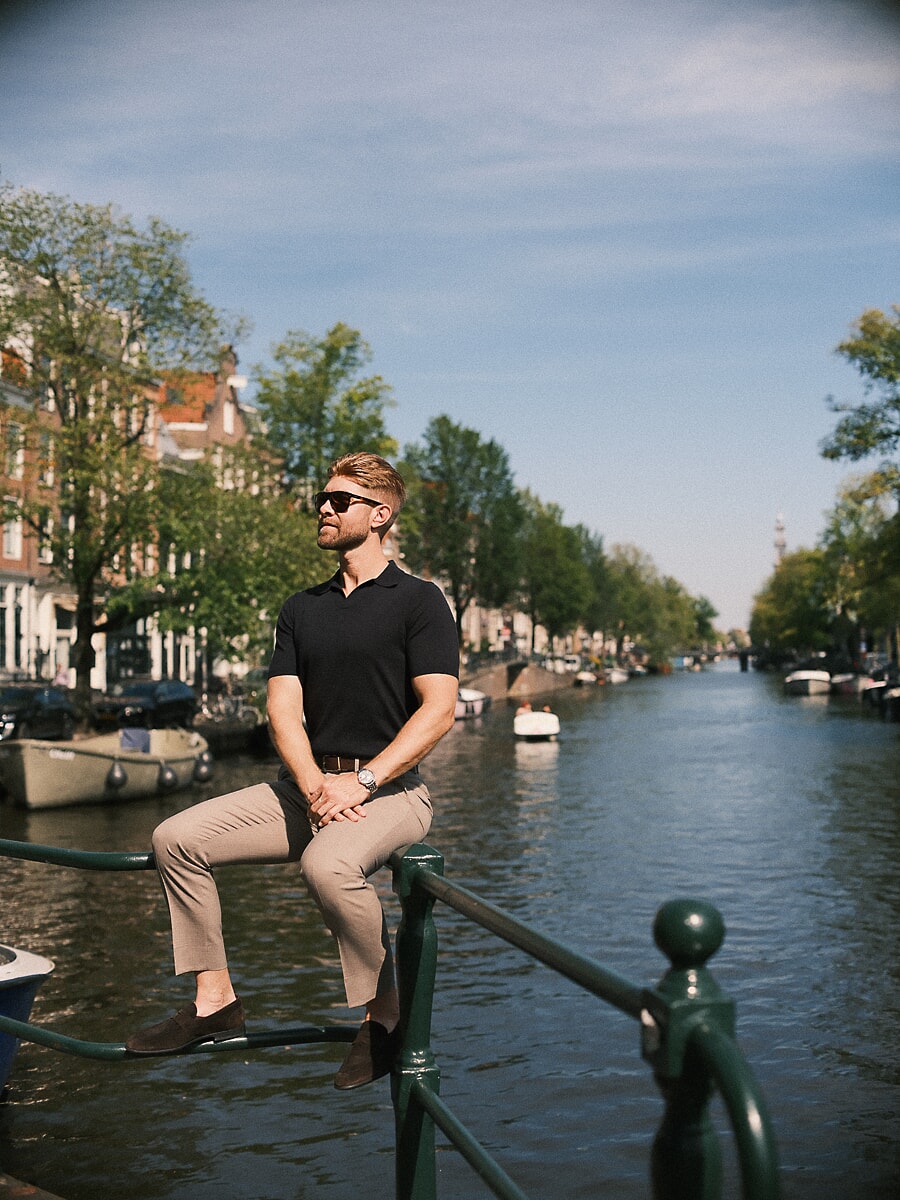 Man met zonnebril zit op een groene reling langs de gracht met wijds uitzicht over het water, een lifestyleportret in Amsterdam