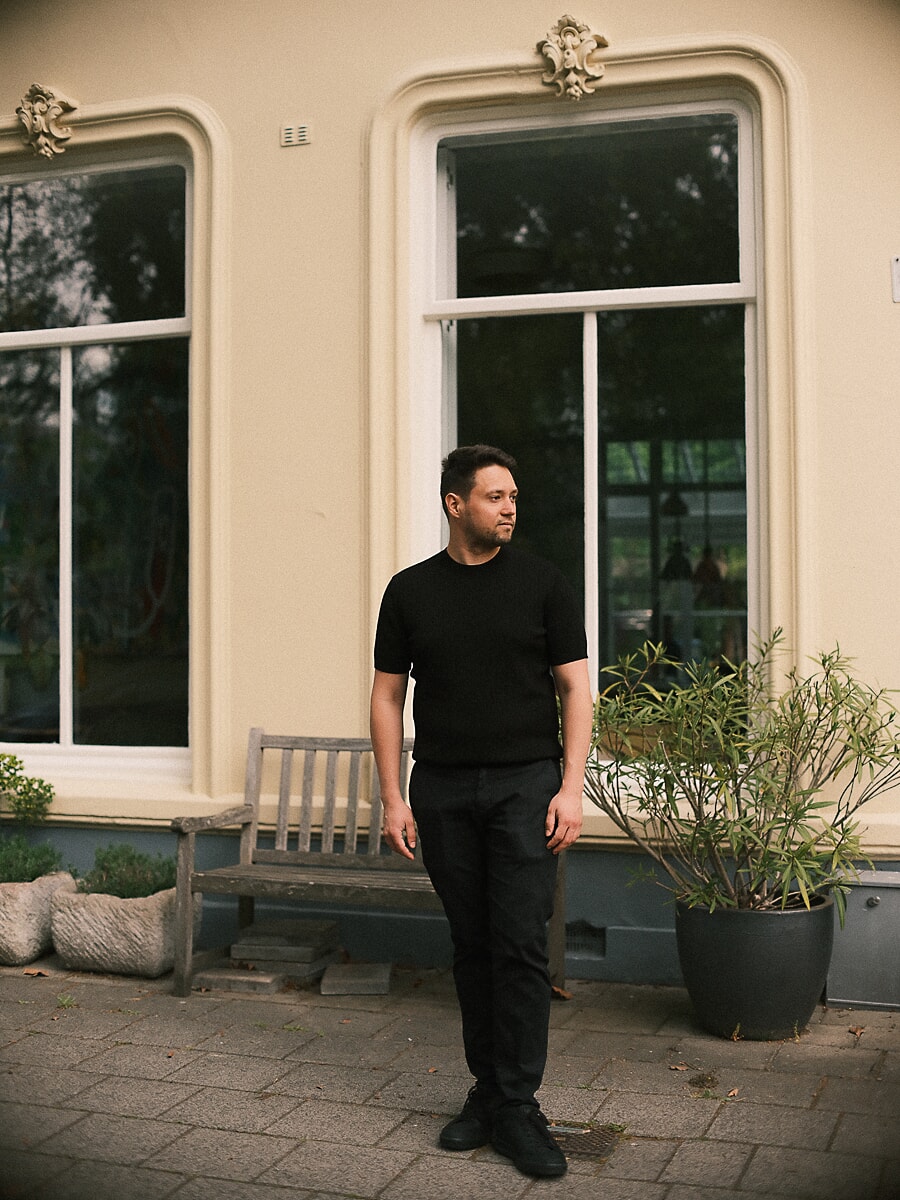 Man in zwart T-shirt voor een crèmekleurige villa met hoge sierlijke ramen, met een bankje en plantenbak naast hem