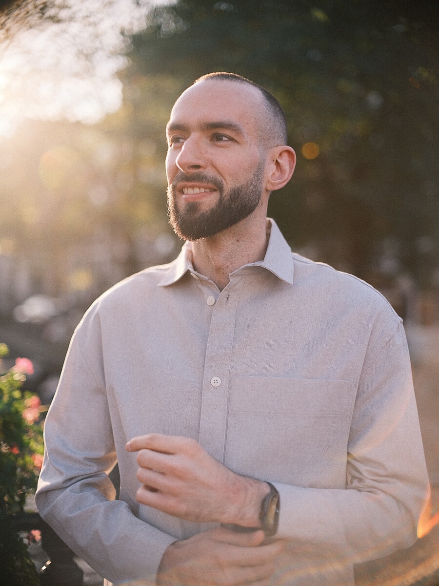 Close-up van een man met baard in een licht linnen overhemd, met sterke zonnegloed van links langs roze bloemen