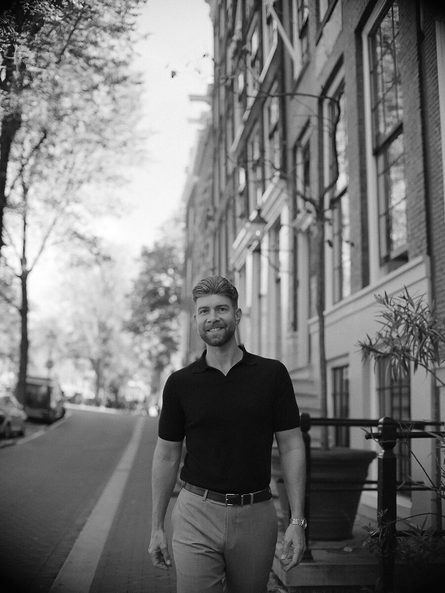 Man in donkere polo loopt door een lommerrijke straat langs een gracht, een breed zwart-wit beeld uit zijn portretshoot