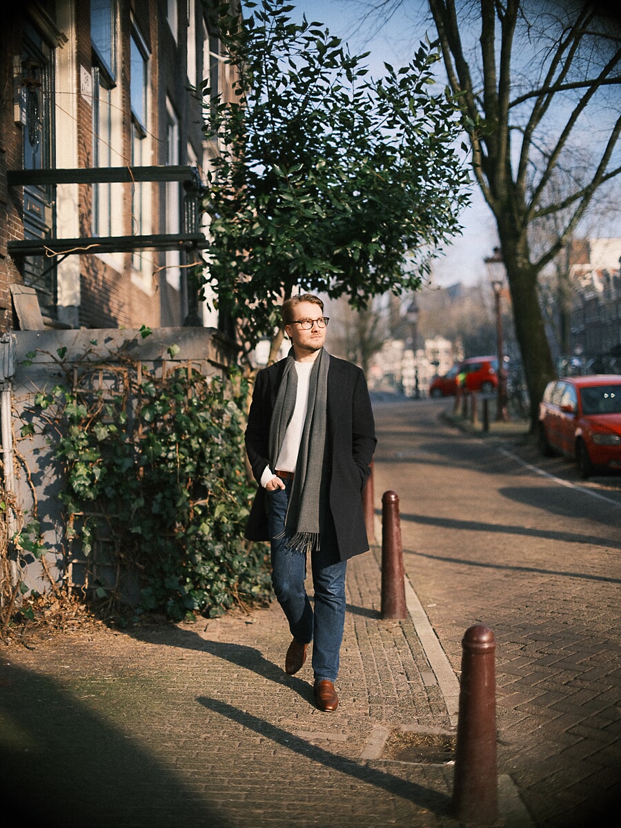 Man met bril en lange zwarte jas loopt door een zonnige straat langs een gracht in de winter, een ontspannen datingportret