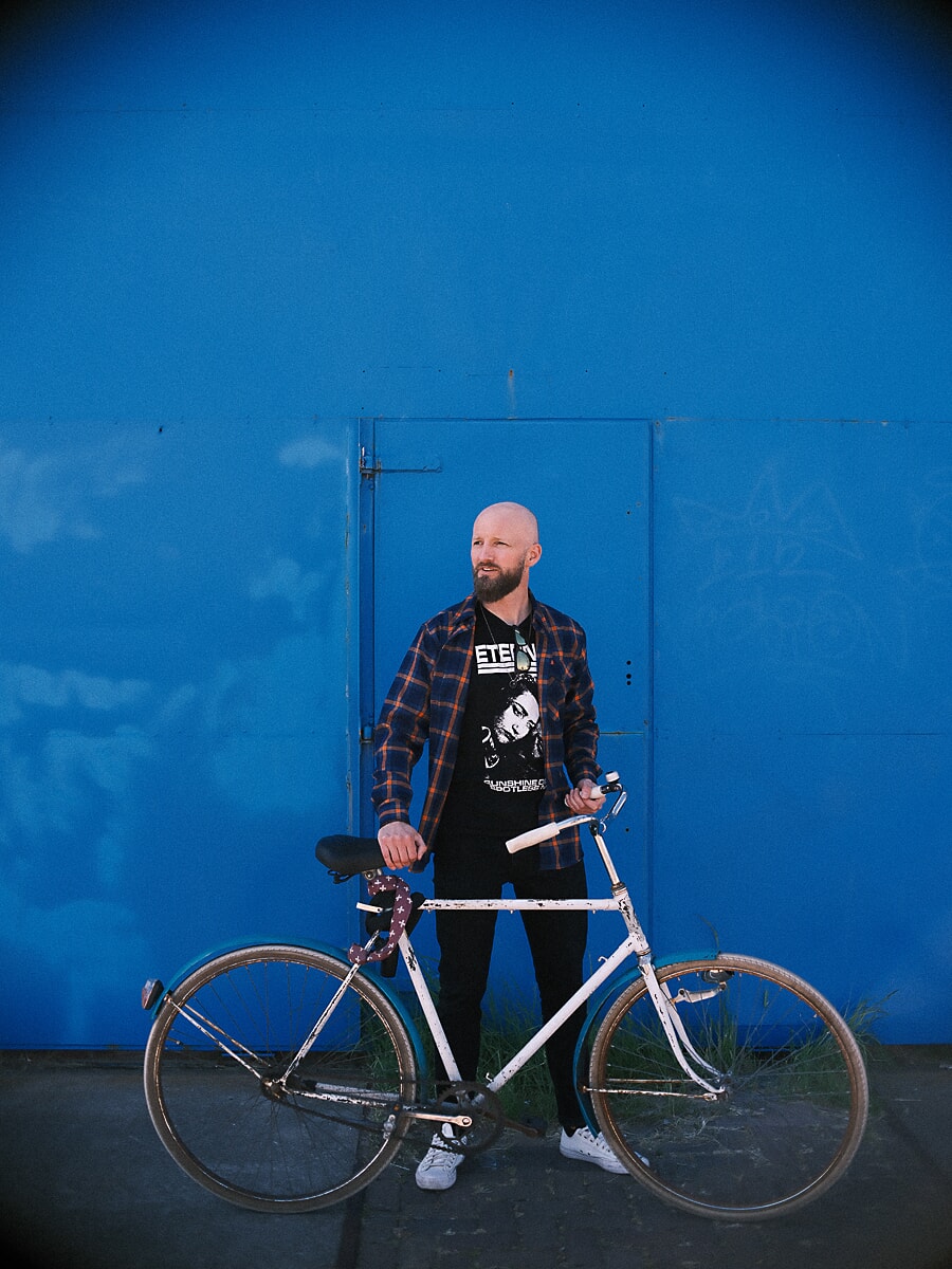 Man in geruit overhemd achter een witte vintage fiets tegen een felblauwe muur die het hele beeld achter hem vult
