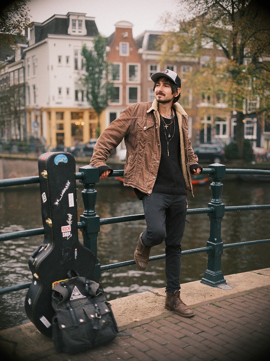 Man met pet en corduroy jack leunt tegen een brugleuning met zijn gitaarkoffer naast zich, een datingportret in Amsterdam