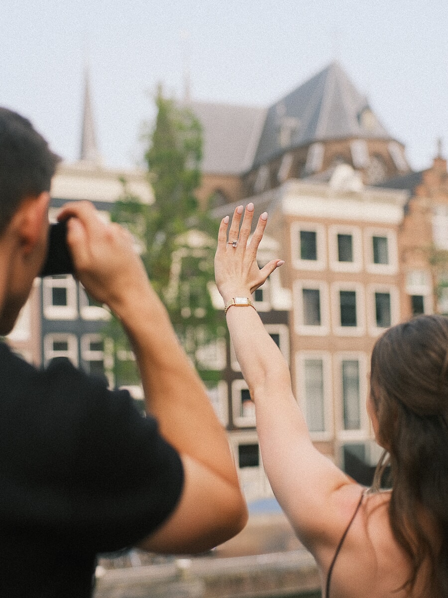 Pas verloofde vrouw toont haar ring terwijl haar partner haar fotografeert bij Amsterdamse grachtenpanden