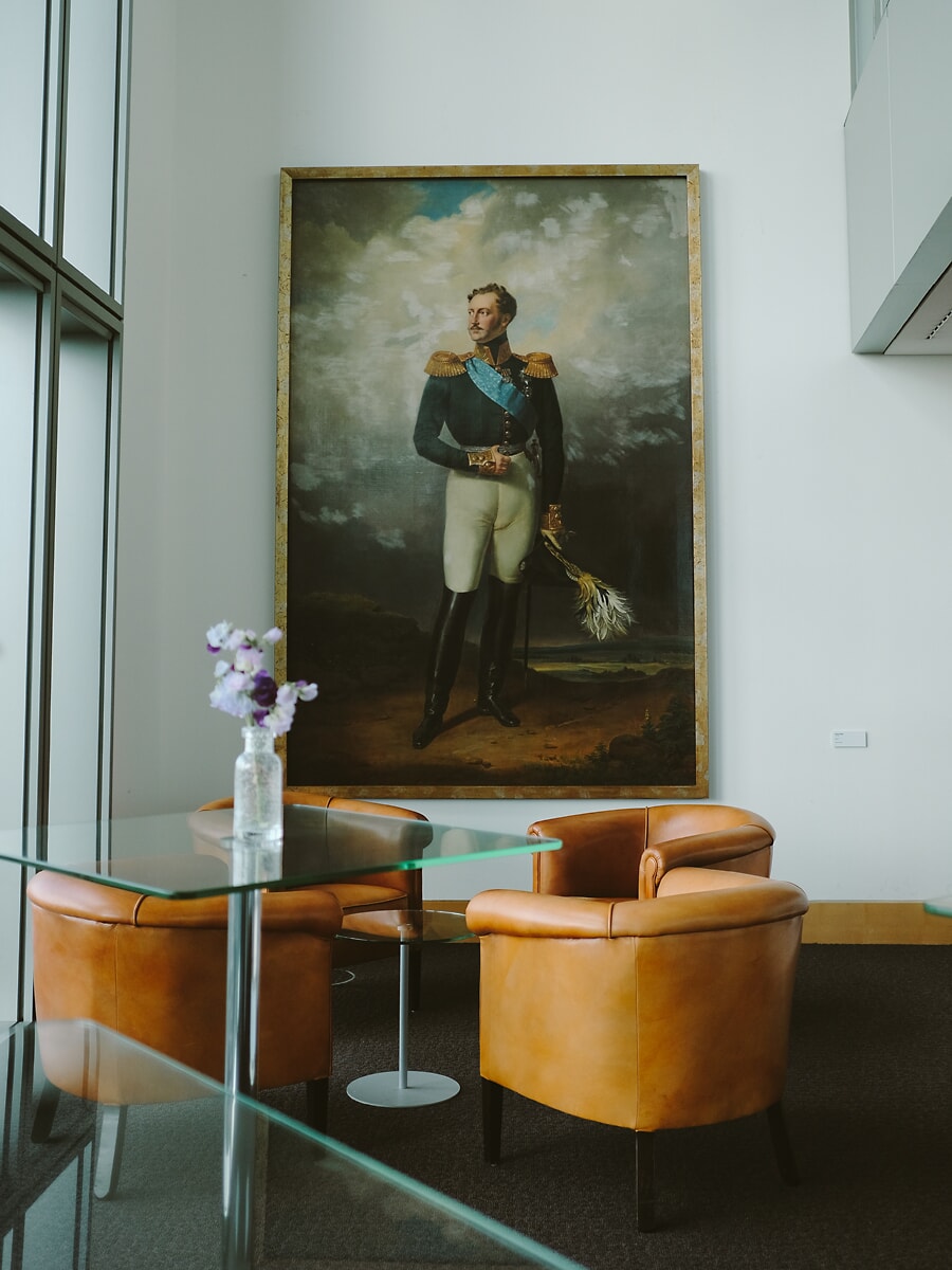 Loungehoek met cognackleurige leren fauteuils en glazen tafels onder een groot klassiek portret, een kantoorinterieur in Amsterdam