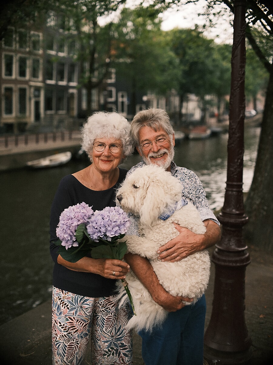 Ouder stel lacht aan een Amsterdamse gracht, zij met lila hortensia's, hij met hun grote krulharige hond op de arm