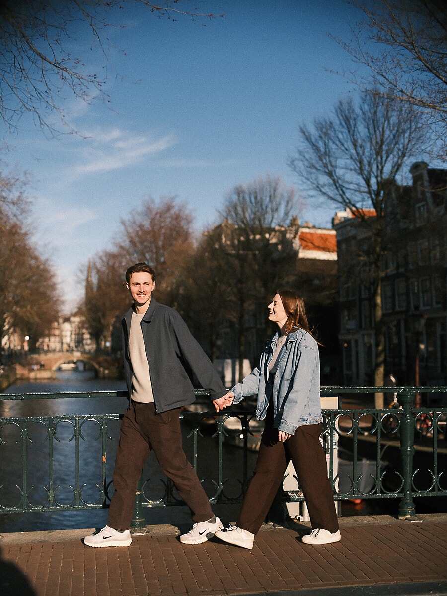 Stel loopt hand in hand over een Amsterdamse grachtbrug op een heldere winterdag, een pre-wedding koppelportret