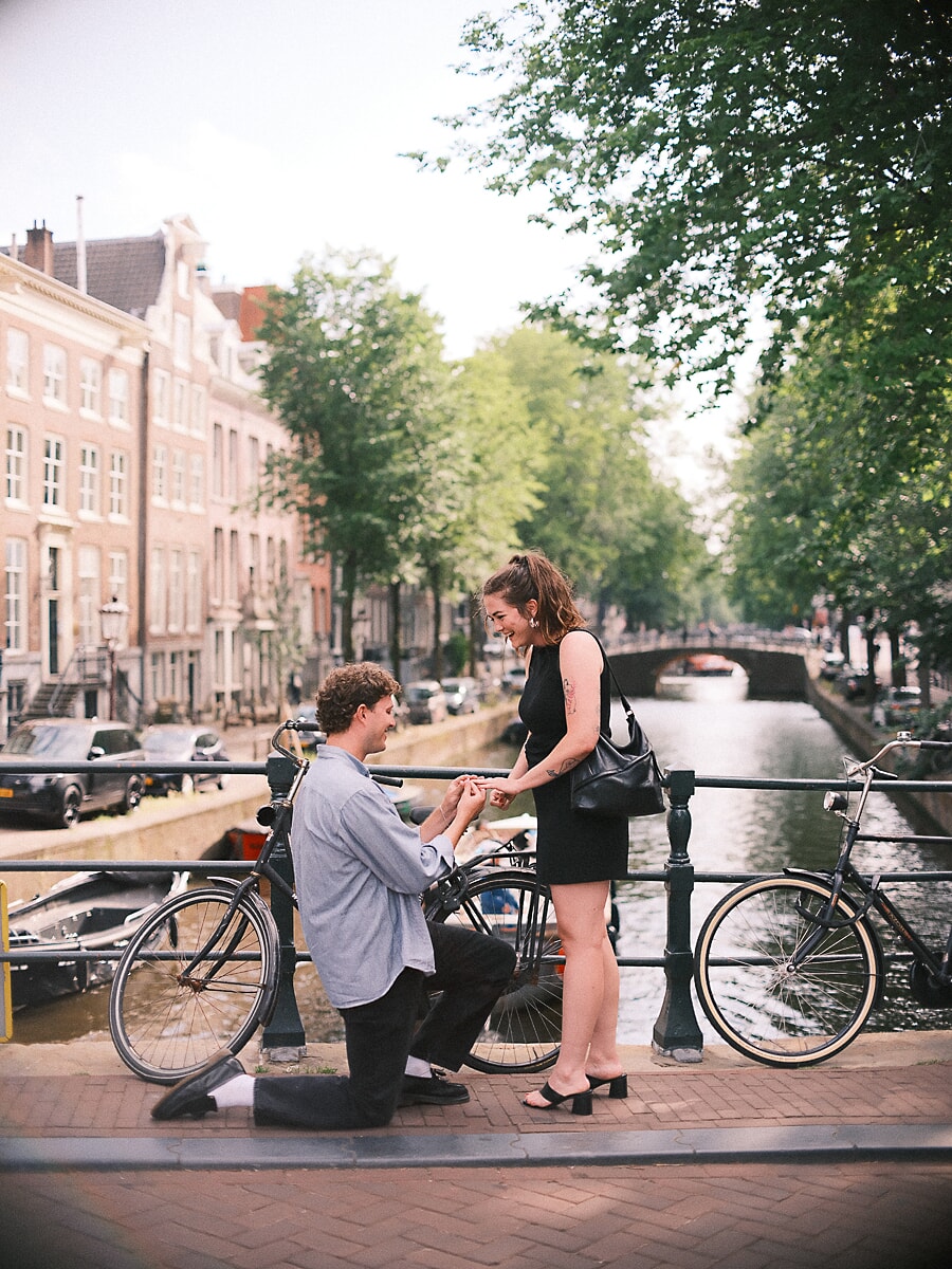 Verrassingsaanzoek tussen geparkeerde fietsen op een Amsterdamse grachtbrug, terwijl zijn partner lachend reageert