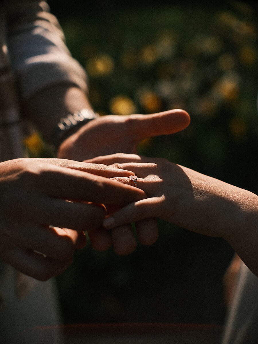 Twee handen komen samen terwijl de verlovingsring om haar vinger wordt geschoven, met zonverlichte bloemen erachter