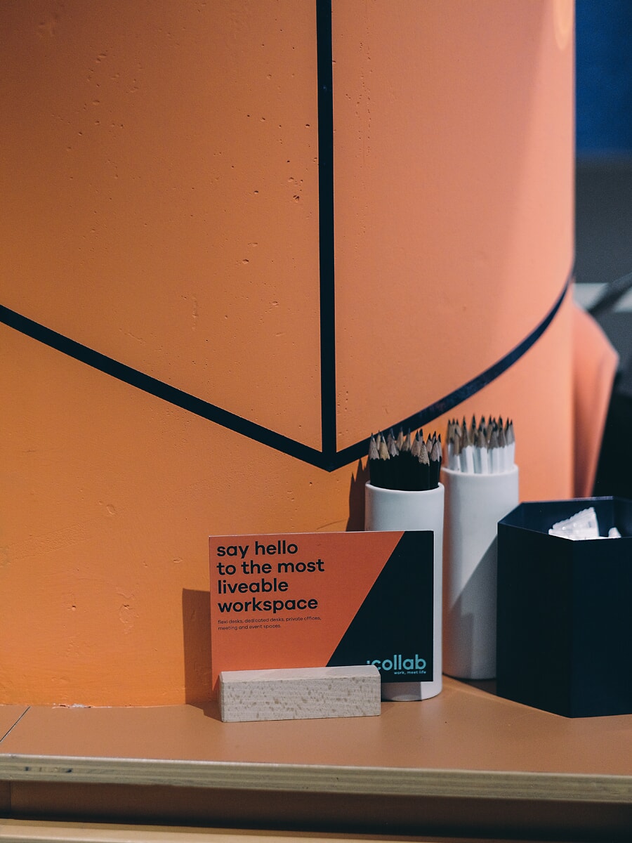 Detail van een receptiebalie: een klein gedrukt kaartje tussen potten met potloden tegen een oranje geometrische wand