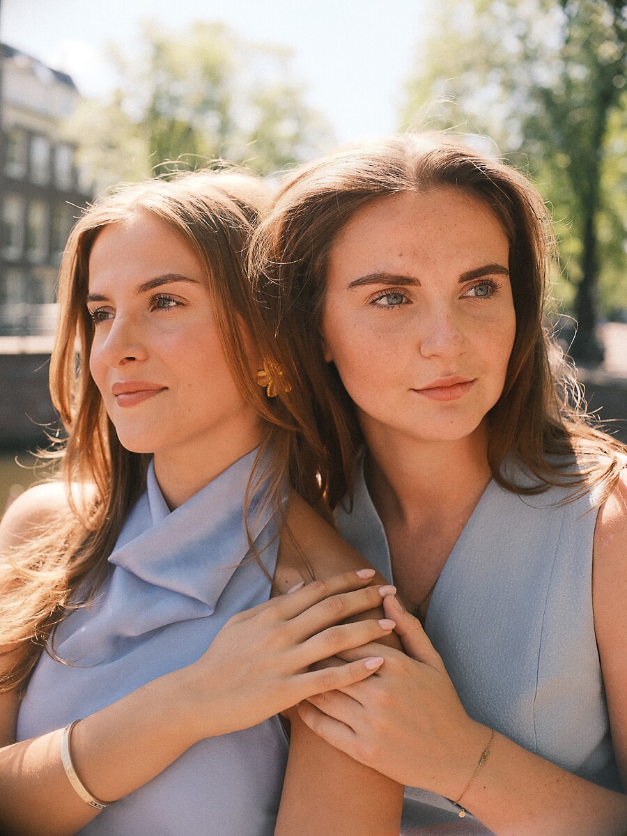 Twee zussen in lichtblauw dicht tegen elkaar in warm tegenlicht, de een leunt tegen de schouder van de ander, handen ineen