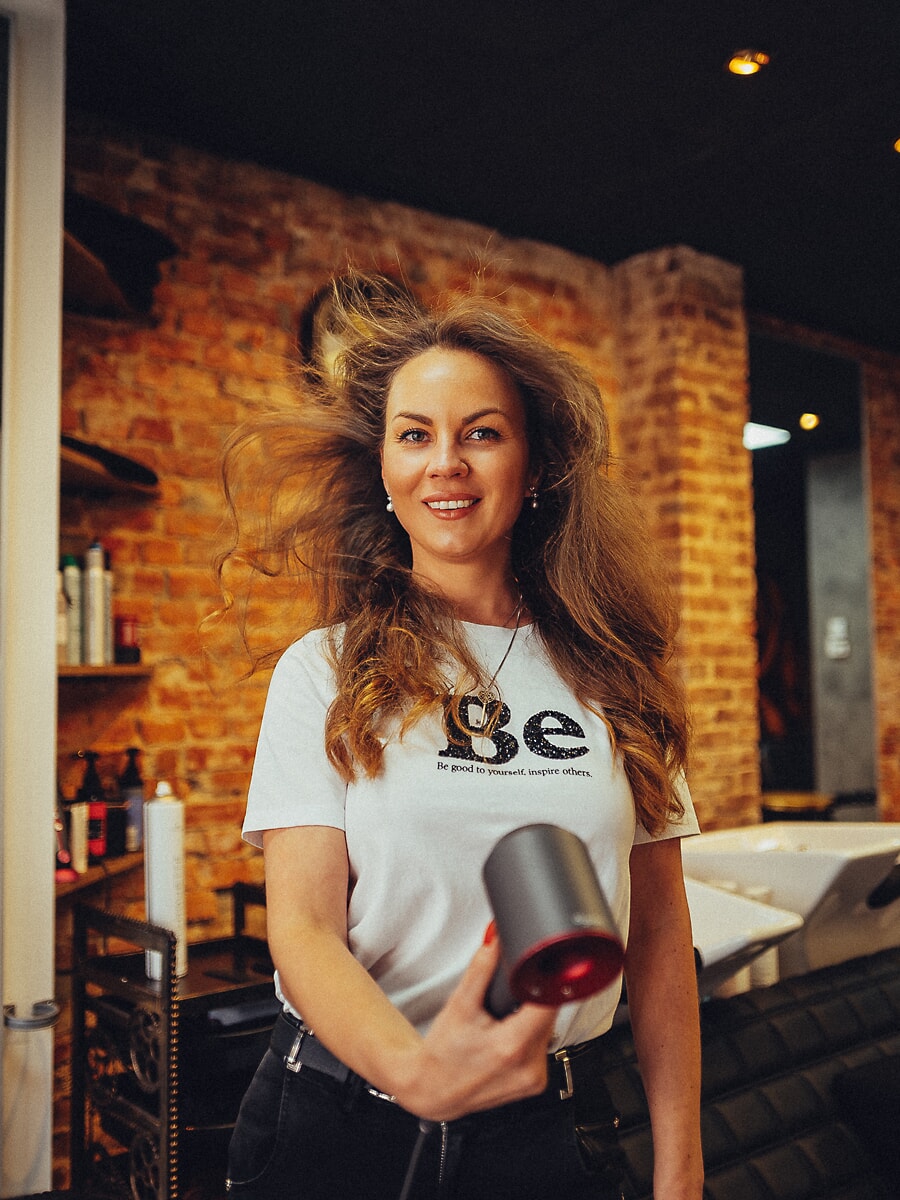 Stylist met een föhn terwijl de luchtstroom haar haar optilt, bakstenen muur en productschappen achter haar, productfotografie in Amsterdam