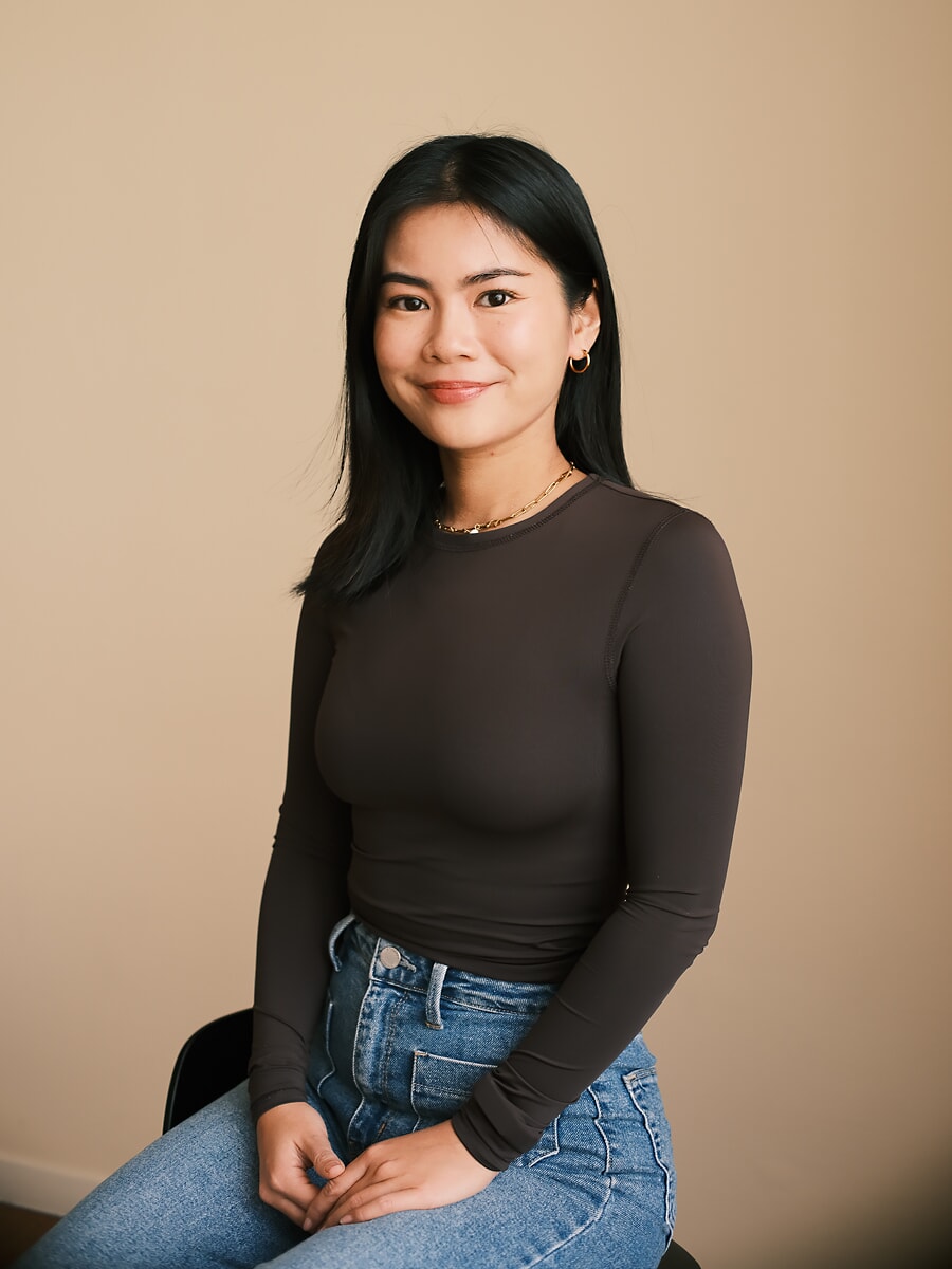 Collega in een bruine longsleeve en jeans zit met haar handen in haar schoot tegen een effen muur