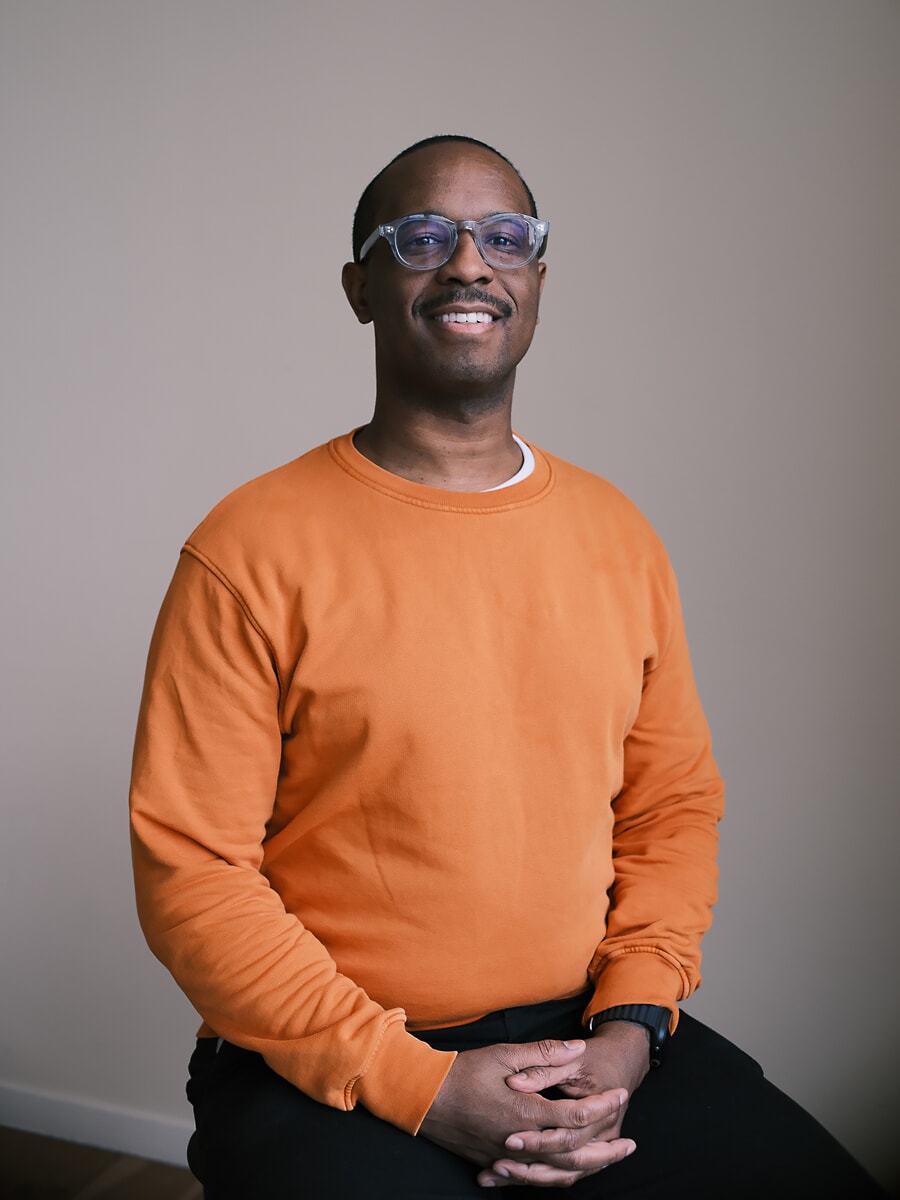 Collega in een oranje sweater met een transparante bril zit met gevouwen handen en lacht naar de camera