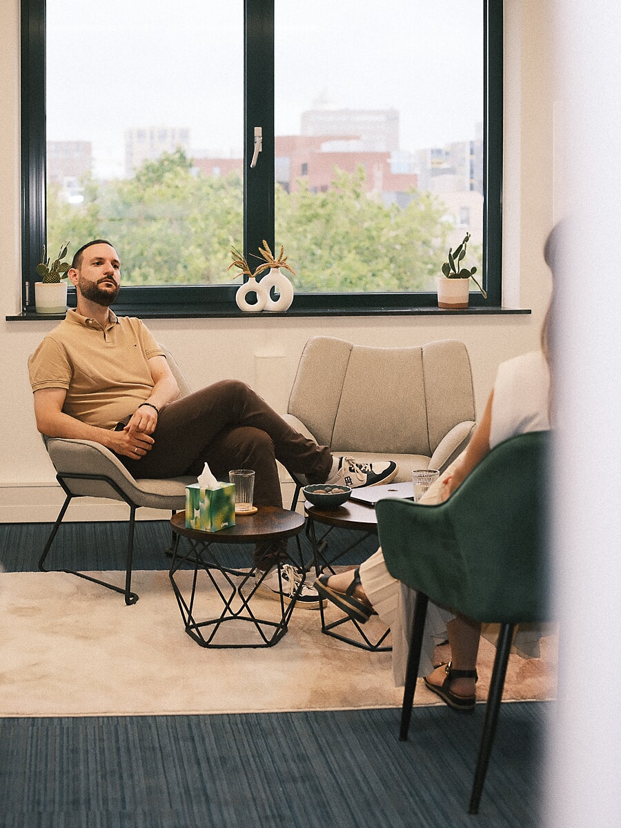 Therapeut in een beige polo luistert vanuit een lage stoel bij een breed raam, met daken erachter, een brandingshoot in Amsterdam