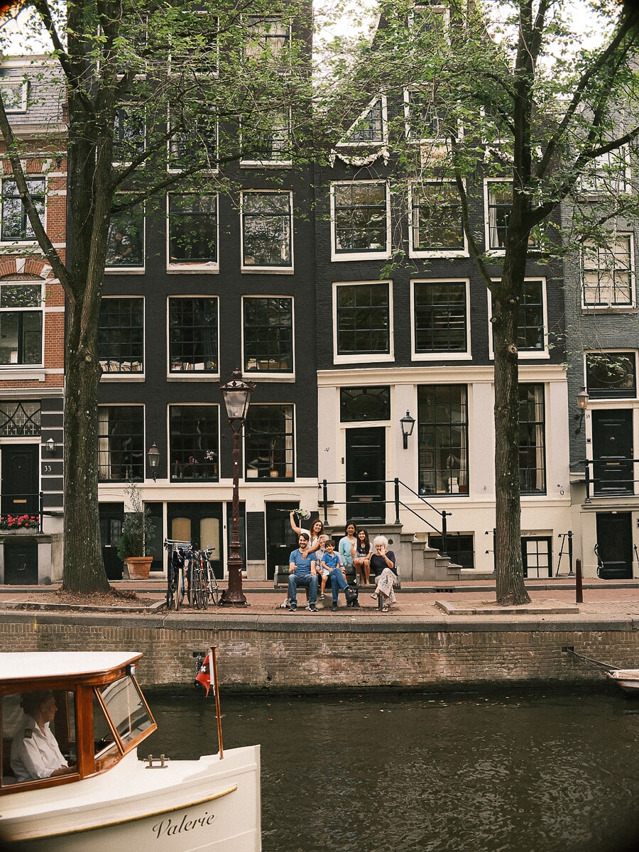 Drie generaties van een familie samen op de stoep van een Amsterdams grachtenpand, gezien over het water terwijl een salonboot voorbijvaart