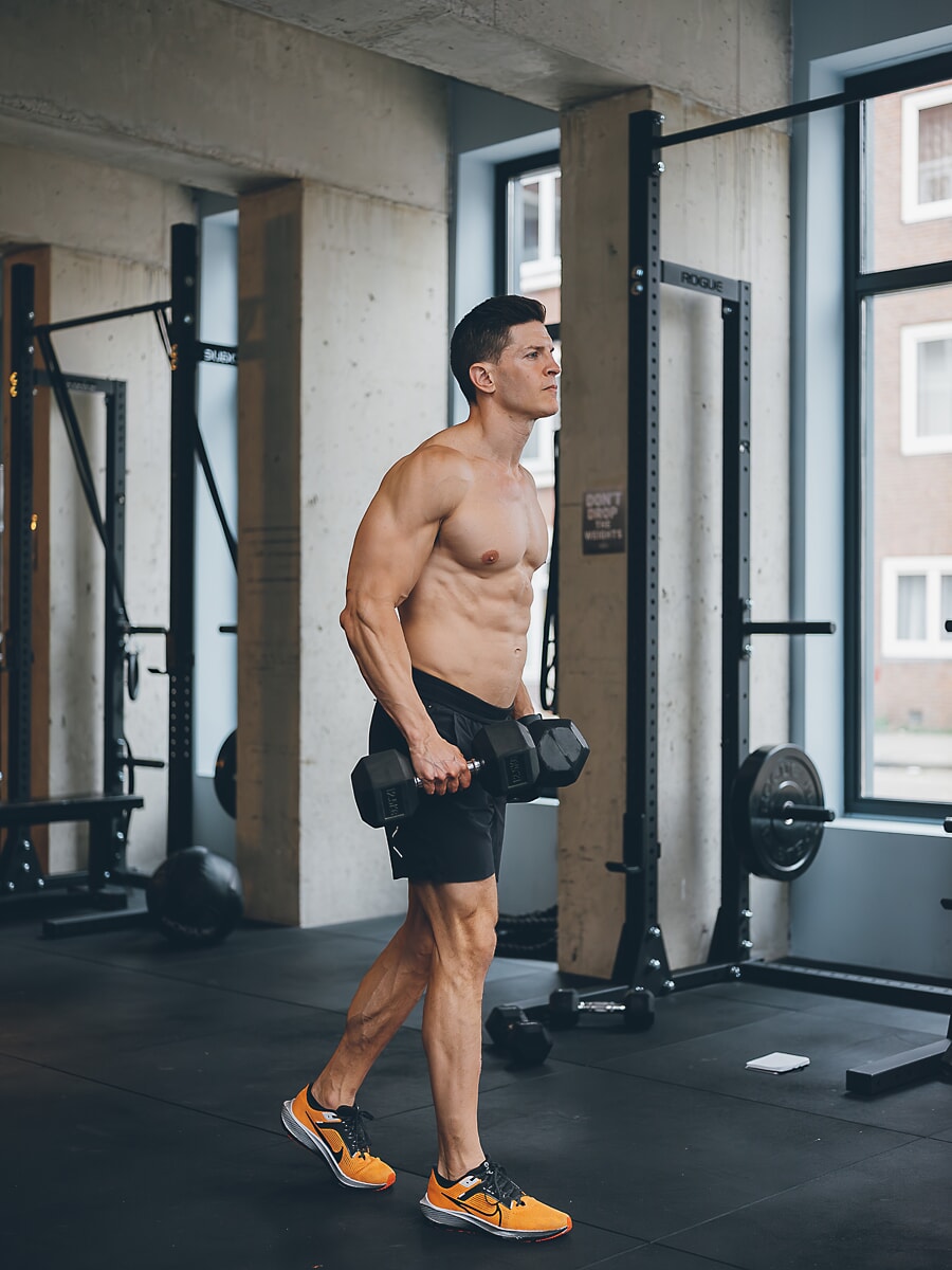 Trainer loopt met twee dumbbells over de betonnen sportschoolvloer, squatrekken en hoge ramen achter hem
