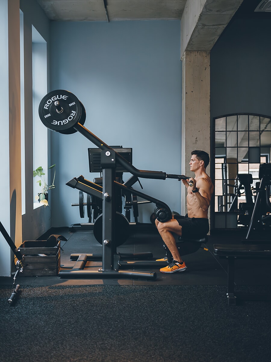 Trainer werkt op een machine met gewichtsschijven in een betonnen sportschool, tegen een lichtblauwe muur, brandingfotografie in Amsterdam