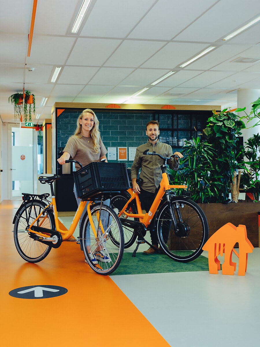 Twee medewerkers bij twee oranje e-bikes in een licht kantoor, met oranje vloermarkeringen, een bedrijfsfotoshoot in Amsterdam
