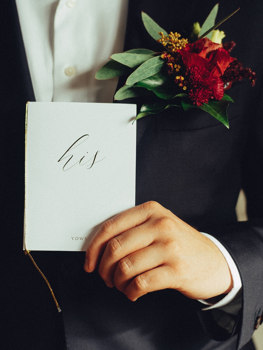 Wedding Ceremony SP10 Een 'his'-gelofteboekje in de hand van de bruidegom met een donkerrode corsage op zijn revers, een intiem ceremoniedetail uit de bruiloftsfotosessie