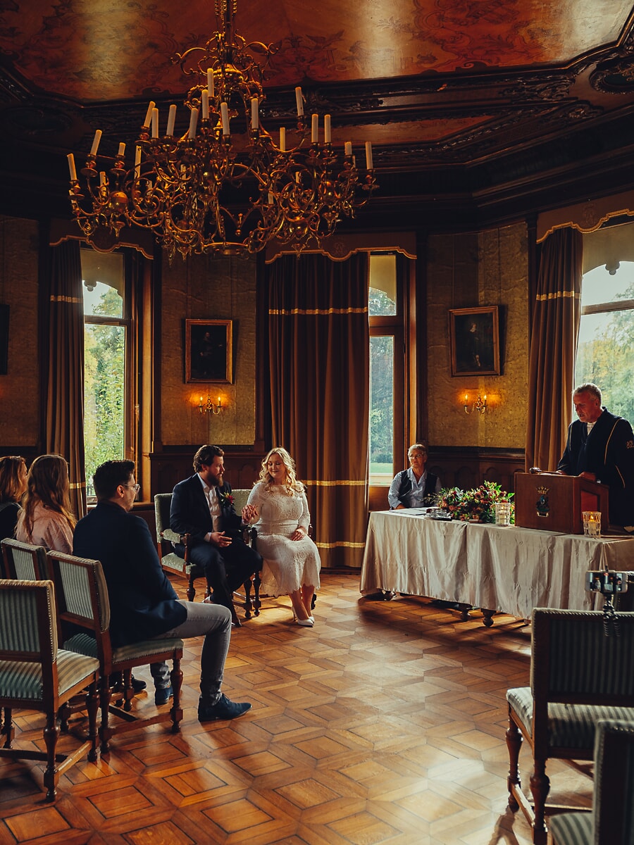 Wedding Ceremony SP13 Burgerlijk huwelijk in een zaal met kroonluchter, het bruidspaar zit voor de ambtenaar, vastgelegd door een trouwfotograaf in Amsterdam