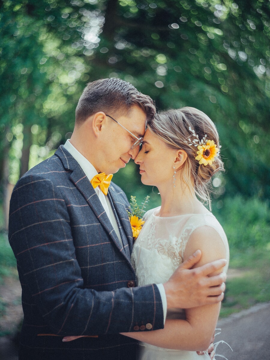 Wedding Ceremony SP14 Bruidegom in een geruit pak leunt met zijn voorhoofd tegen dat van zijn bruid op een groen bospad, een zonnebloem in haar haar, een teder trouwportret