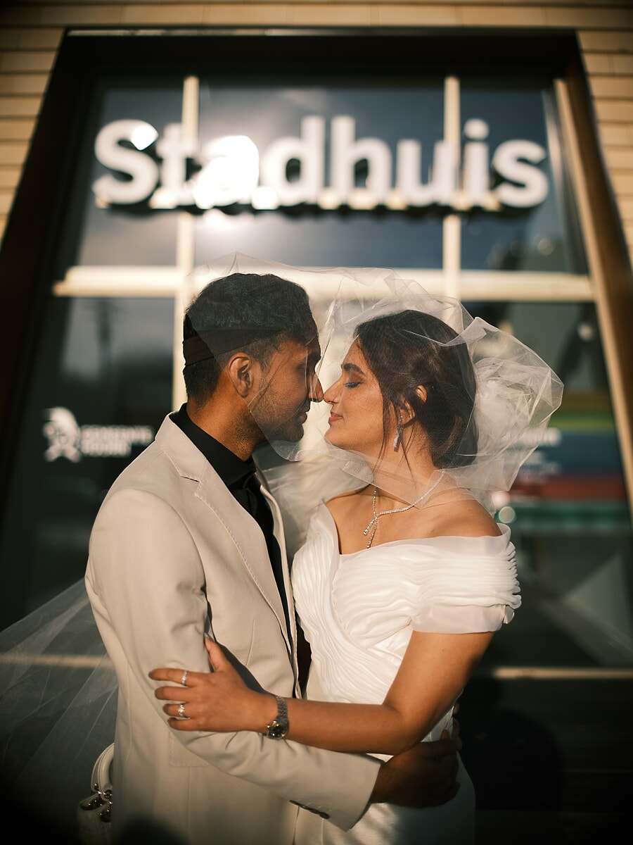 Wedding Ceremony SP2 Pasgetrouwden raken elkaar met het voorhoofd aan onder de sluier voor een verlicht stadhuis in de schemering, een teder moment uit hun trouwshoot