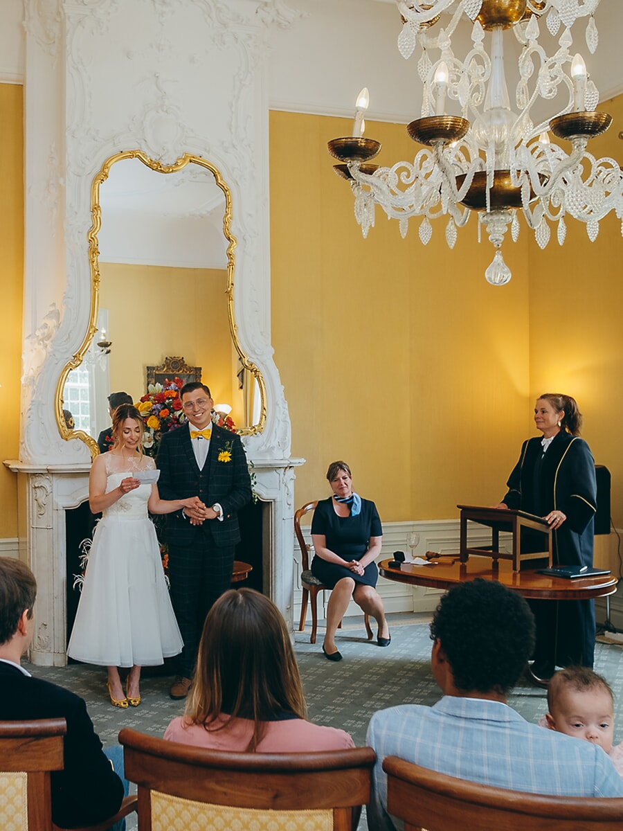 Wedding Ceremony SP23 Met haar geloften in de hand staat de bruid voor de ambtenaar in een verguld stadhuis, gasten kijken toe, een spontane ceremoniefoto